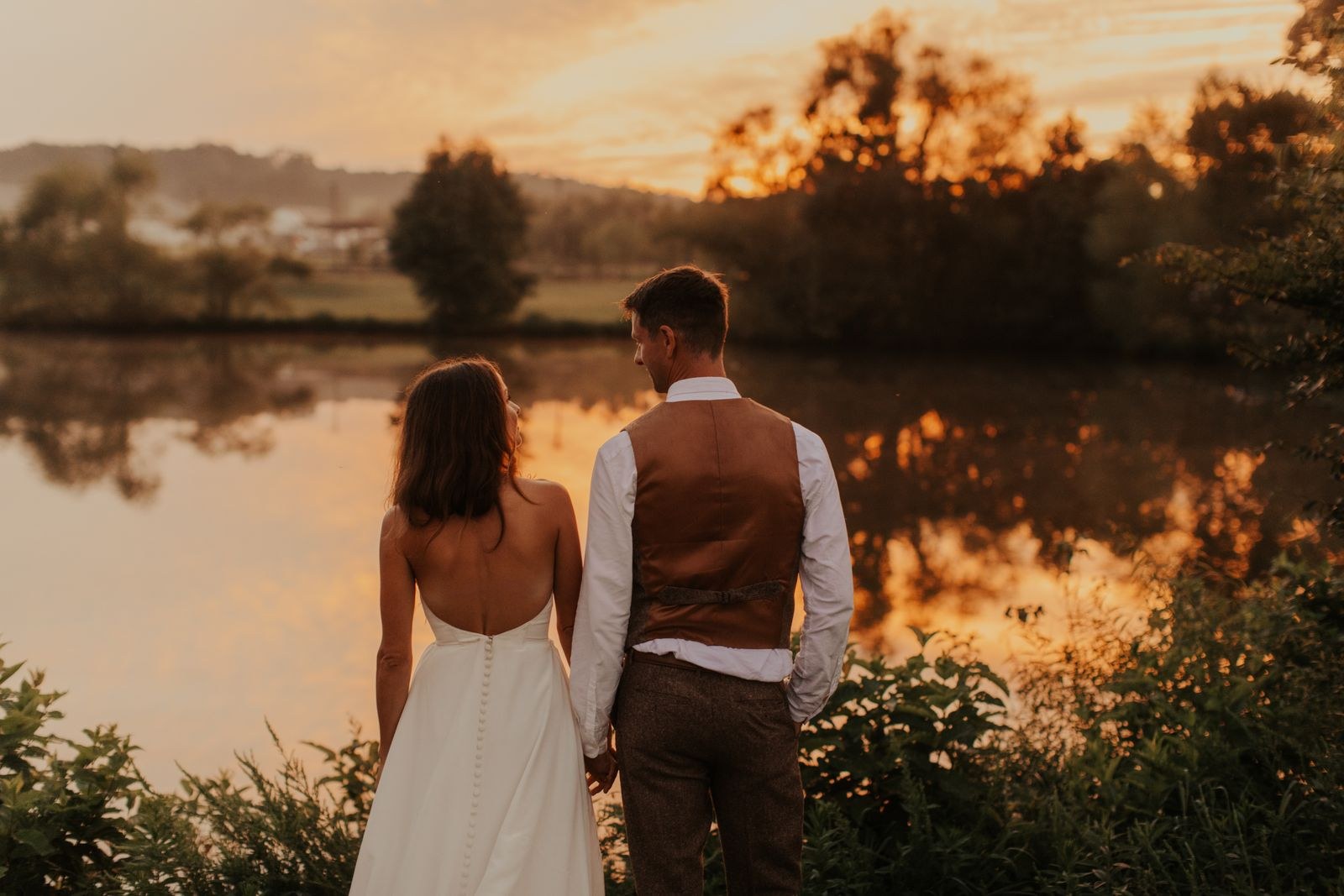 Couple standing hand in hand facing the pond at sunset